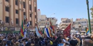 Una folla radunata protesta davanti a un edificio in Siria, issando varie bandiere.