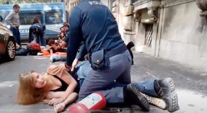 una donna è per terra di fianco, sul marciapiede, con un agente di polizia che la blocca a terra. Dietro si intravedono altri agenti con altre persone sdraiate e tenute in stato di fermo da altri agenti davanti a una camionetta della polizia