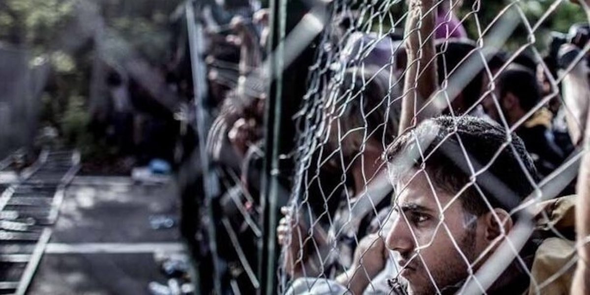 Una persona con il viso dietro una rete metallica al confine orientale dell'Europa. L'Europa respinge i migranti pur avendone bisogno.