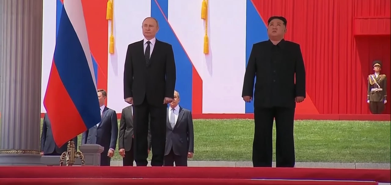 Vladimir Putin e Kim Jong Un in posa, dietro di loro alcuni funzionari della Corea del Nord e un militare sullo sfondo. Accanto a Putin c'è una bandiera della Russia