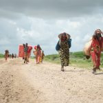 Donne, che camminano con ciò che riescono a portare, diretto verso un campo per sfollati eretto accanto a una base militare per le vittime delle forti piogge a causa della crisi climatica.