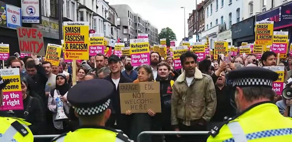 Un gruppo nel Regno Unito di manifestanti venuti a difendere le proprie comunità contro gli attacchi razzisti degli scorsi giorni. di fronte al cordone di poliziotti, reggono cartelloni con varie scritte: "smash fascism and racism" "Farage and EDL not welcome here" "refugee welcome" "stop the far-right"