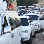 una fila di auto in fuga ferme nel traffico - un gruppo di tre uomini sul ciglio della strada guarda le auto. Nell'abitacolo della prima si intravedono dei bambini.