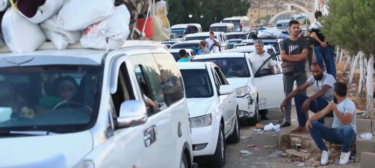 una fila di auto in fuga ferme nel traffico - un gruppo di tre uomini sul ciglio della strada guarda le auto. Nell'abitacolo della prima si intravedono dei bambini.