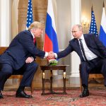 Donald Trump (a sinistra, seduto) e Vladimir Putin (a destra, seduto) si stringono la mano. In mezzo a loro un tavolino. La stanza ha un tappeto con motivi orientali, alle spalle dei due invece bandiere degli Stati Uniti e della Federazione russa