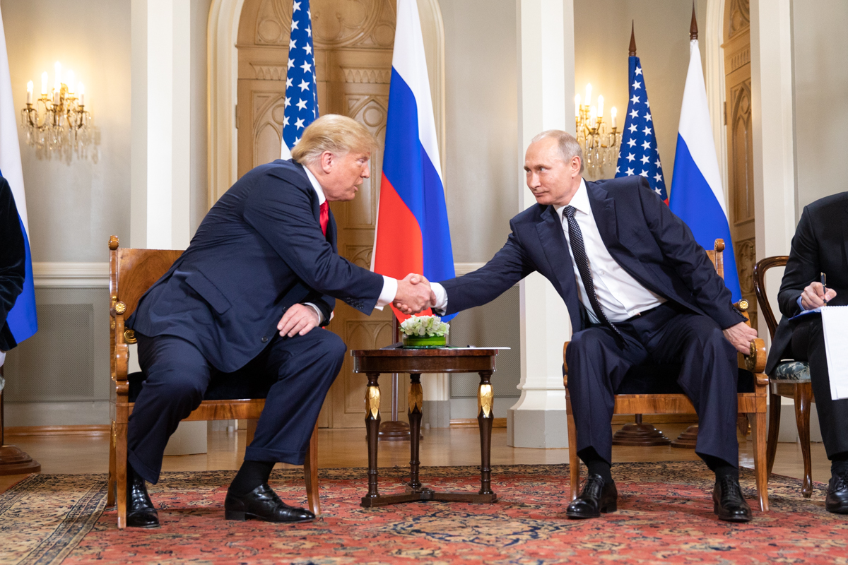 Donald Trump (a sinistra, seduto) e Vladimir Putin (a destra, seduto) si stringono la mano. In mezzo a loro un tavolino. La stanza ha un tappeto con motivi orientali, alle spalle dei due invece bandiere degli Stati Uniti e della Federazione russa