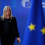 La presidente del Consiglio italiana Giorgia Meloni passeggia con aria seria. Sullo sfondo le bandiere dell'Unione Europea