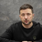 Volodymyr Zelensky seduto a un tavolo con le mani incrociate. Alle sue spalle, sulla sinistra del video, una bandiera dell'Ucraina