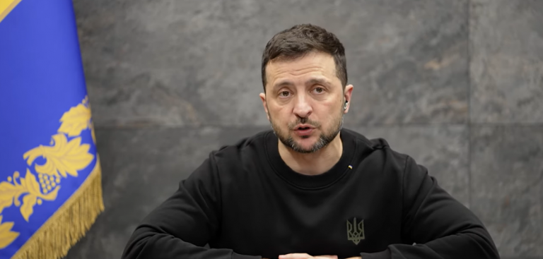 Volodymyr Zelensky seduto a un tavolo con le mani incrociate. Alle sue spalle, sulla sinistra del video, una bandiera dell'Ucraina