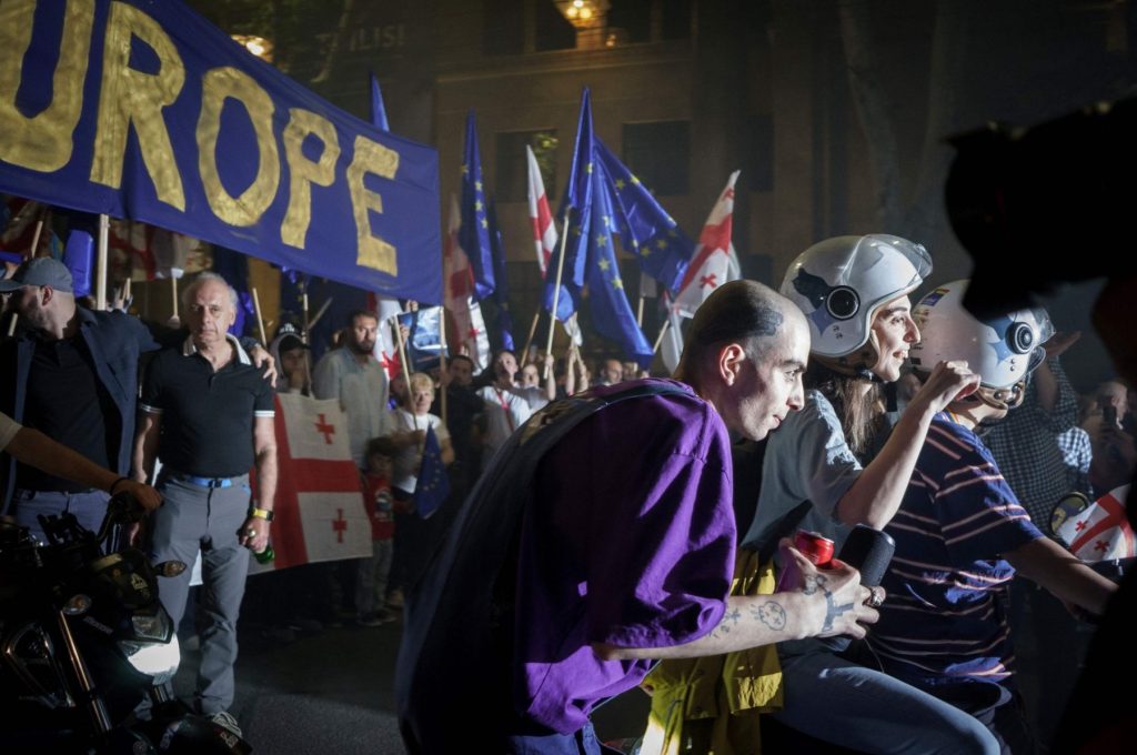 Gruppo di manifestanti georgiani. Davanti alla file una coppia su uno scooter, e accanto a loro un manifestanti chino in avanti, con due lattine in mano.
Dietro, striscioni con scritto "Europe", bandiere dell'Unione Europea e della Georgia.