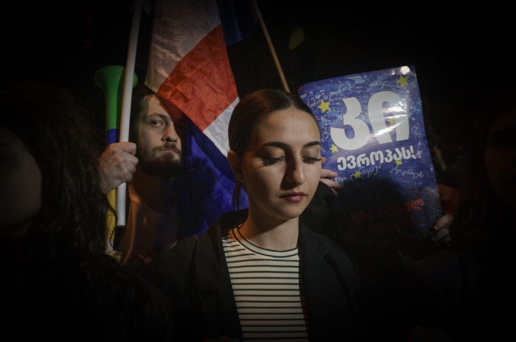 una giovane manifestante georgiana con lo sguardo rivolto verso il basso. Dietro di lei, in penombra, si intravedono altri manifestanti. Uno è un giovane con la barba, e imbraccia una bandiera del paese.