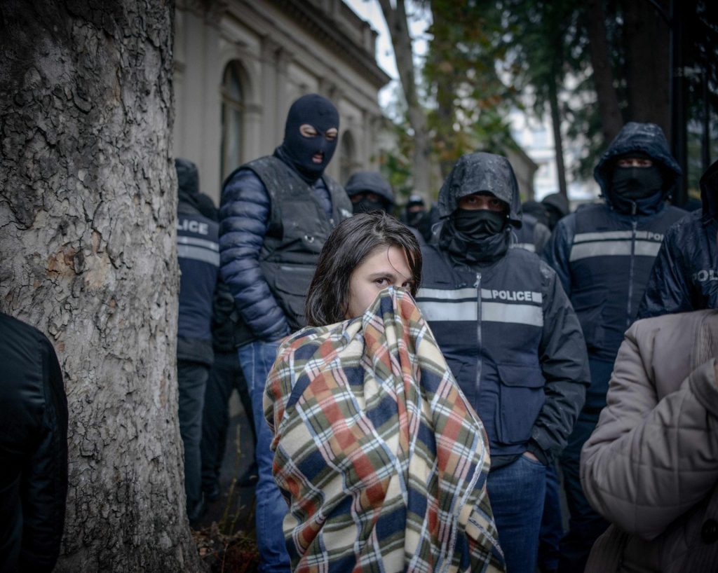 Una manifestante avvolta da una coperta si copre il volto. Dietro di lei un gruppo di poliziotti incappucciati e/o con passamontagna.