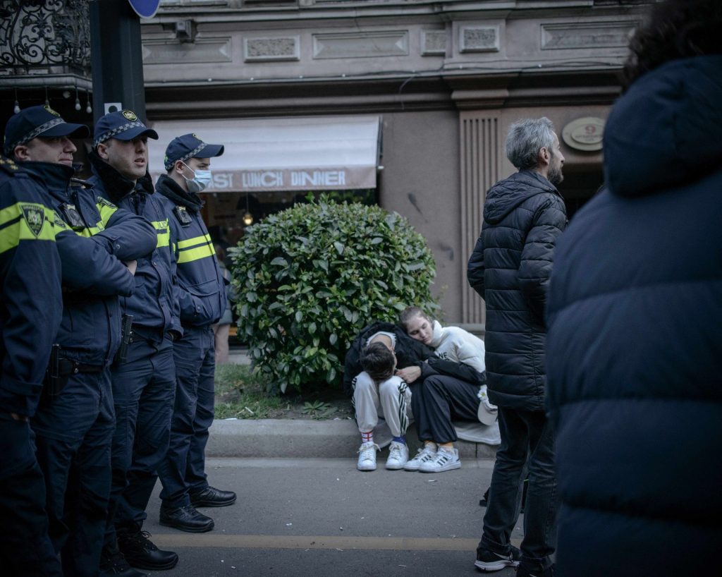 Una coppia seduta sull'asfalto, assonnata. Una persona ha la testa appoggiata sulle gamba, l'altra l'abbraccia e tiene la testa appoggiata sulla sua spalla. I due sono fotografati tra due gruppi di poliziotti: una fila di poliziotti in piedi sulla strada, e un gruppo di manifestanti davanti a loro.