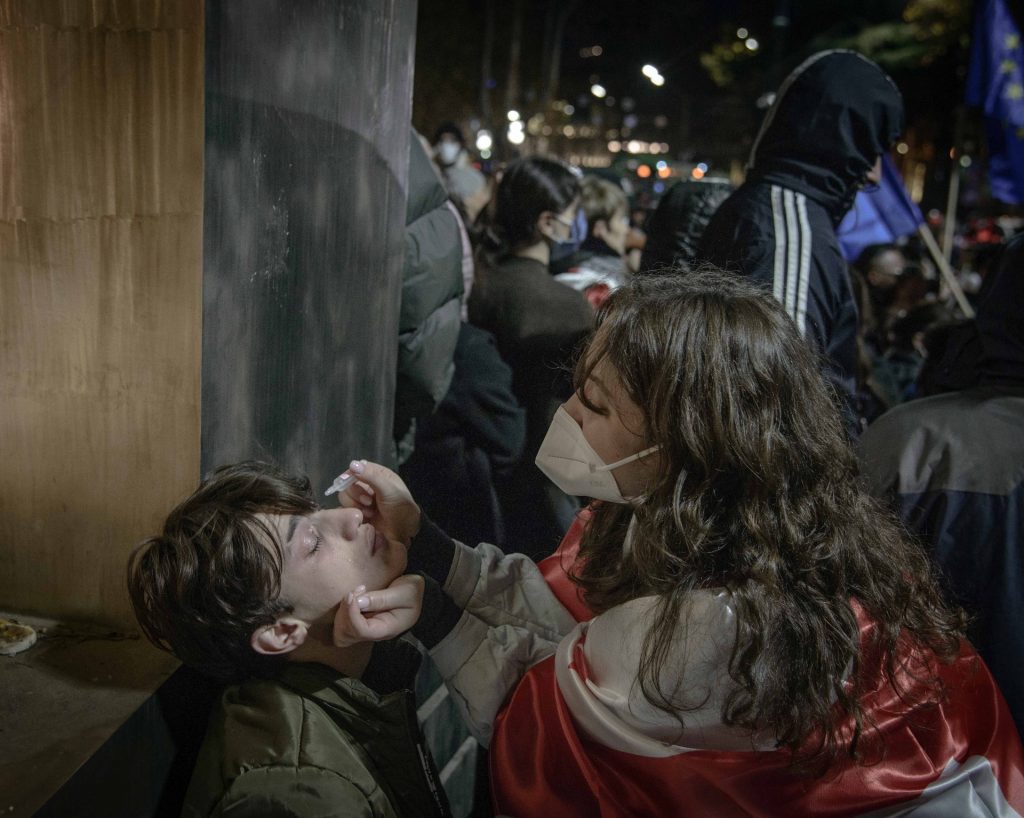 Un ragazzo colpito dai lacrimogeni è seduto e tiene la testa verso l'alto, mentre una giovane con una mascherina medica verso negli occhi soluzione fisiologica. Alle loro spalle un gruppo di manifestanti con il volto coperto, tra cui alcuni con bandiere dell'Unione Europea.