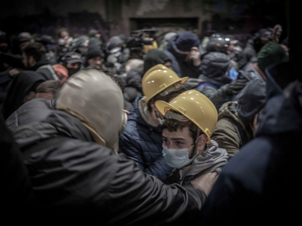 In mezzo a un gruppo di manifestanti, un giovane con elmetto e mascherina medica ha gli occhi lucidi ed è spaventato per i lacrimogeni. Un altro uomo, di spalle e di tre quarti, con indosso una maschera antigas sovietica, gli appoggia una mano sulla spalla per tranquillizarlo.