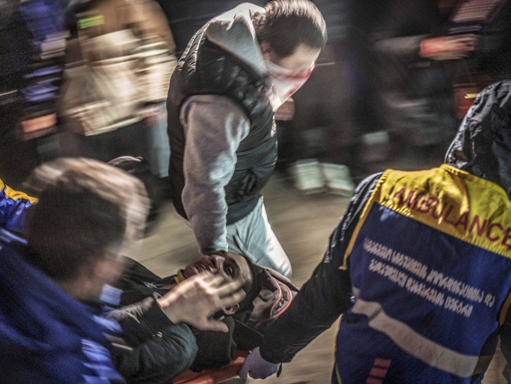 Un manifestante georgiano su una barella è trasportato da due paramedici, davanti e dietro la barella. Un altro manifestanti aiuta il paramedico davanti nel trasporto.