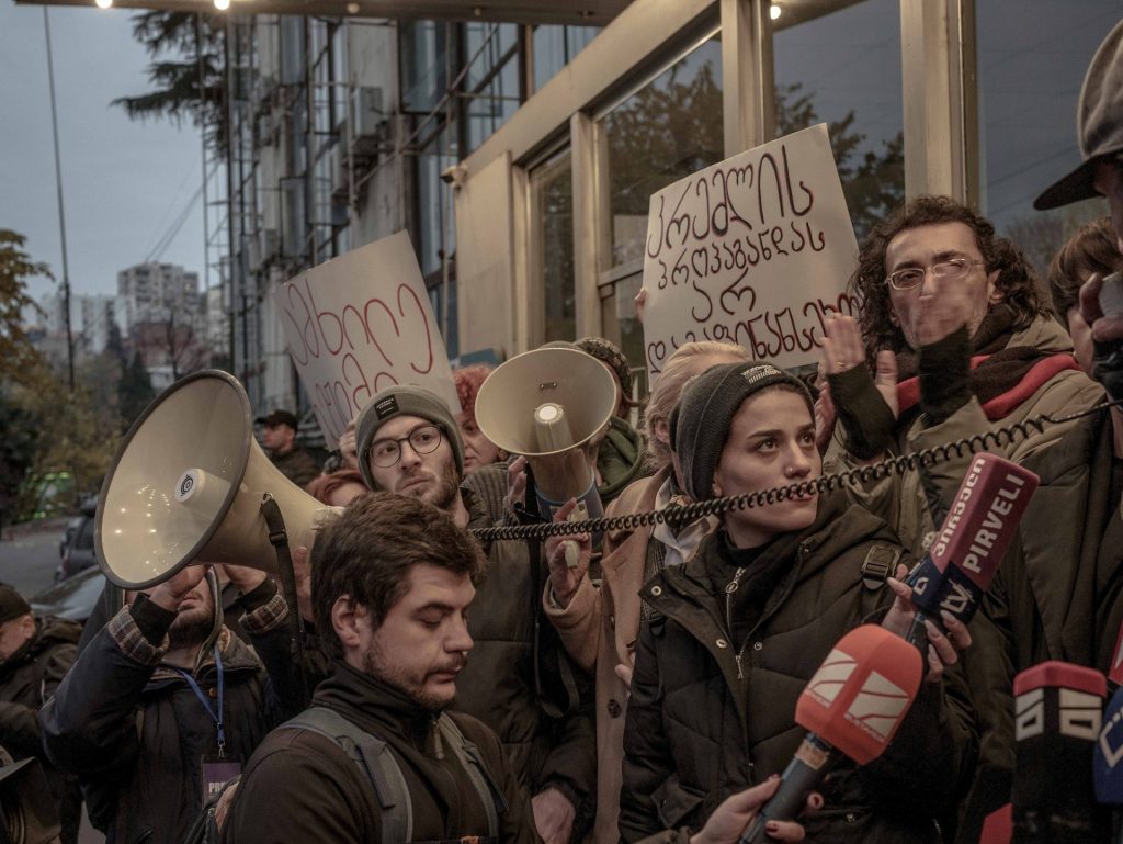 Un gruppo di manifestanti con megafoni e cartelli (in georgiano) è alle spalle di alcuni giornalistiche impugnano microfoni.