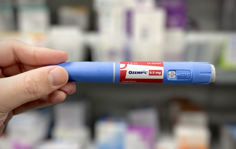 Una mano tiene tra indice e pollice una "penna" da 0,5 di Ozempic. Sullo sfondo, uno scaffale di farmaci