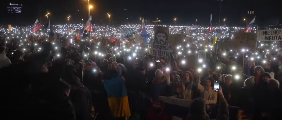 una folla di manifestanti in Slovacchia contro il governo Fico. Alcuni hanno bandiere del paese, altri dell'Ucraina. Il buio della sera è illuminato dagli schermi degli smartphone dei manifestanti, che li impugnano come fiaccole